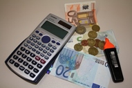 Das Bild zeigt Taschenrechner, Geldscheine und Münzen sowie einen Rotstift